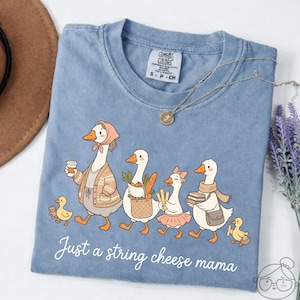 May include: A light blue Comfort Colors t-shirt featuring a whimsical illustration of geese walking in a line. The geese are carrying various items, and the text "Just a string cheese mama" is written in cursive at the bottom.