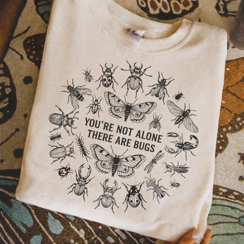 Op de afbeelding: Cr&egrave;mekleurige sweatshirt met een cirkelvormig ontwerp van zwarte insectenillustraties, waaronder vlinders en kevers. De tekst "YOU'RE NOT ALONE THERE ARE BUGS" staat in het midden van het ontwerp.
