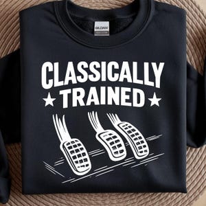 May include: Black sweatshirt with the text "CLASSICALLY TRAINED" in white, with star accents. Below the text are three white illustrations of corn cobs. The sweatshirt is folded on a woven surface.