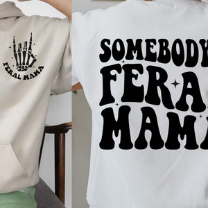 May include: Two sweatshirts with the text "Somebody's Feral Mama" in a retro font. One sweatshirt is beige with a black skeleton hand making a rock and roll sign. The other sweatshirt is white with black text.