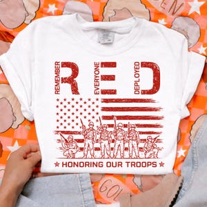 K&ouml;nnte beinhalten: Wei&szlig;es T-Shirt mit roter Grafik. Die Grafik zeigt das Wort "RED" mit den Worten "Remember Everyone Deployed", eine amerikanische Flagge, Soldaten und den Text "Honoring Our Troops."