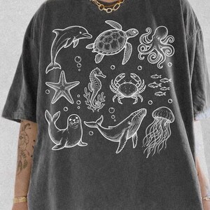 May include: Dark gray t-shirt featuring white illustrations of various sea creatures, including a dolphin, turtle, octopus, starfish, seahorse, crab, fish, seal, whale, and jellyfish. The shirt has a relaxed fit and short sleeves.