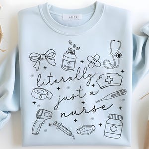 May include: Light blue sweatshirt with the text "literally just a nurse" and medical-themed illustrations in black. The design includes a bow, pills, bandages, a stethoscope, a nurse's hat, a thermometer, a medical mask, a syringe, and a bottle of pills.
