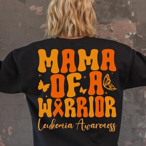 May include: Black sweatshirt with orange and yellow text that reads "Mama of a Warrior" with a butterfly and ribbon design. The text "Leukemia Awareness" is printed below the design.