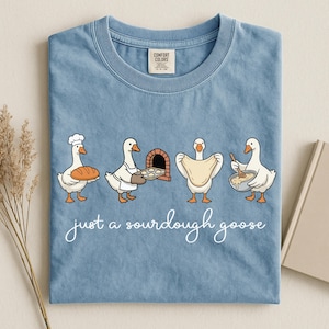 May include: A light blue t-shirt featuring a cartoon of four geese baking sourdough bread. The geese are depicted with a loaf of bread, a tray of rolls, a pizza oven, and a bowl. The text "just a sourdough goose" is written in cursive.