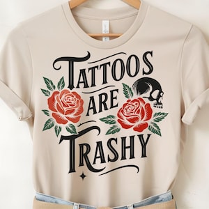 May include: Beige t-shirt with the text "Tattoos are Trashy" in black script, adorned with red roses and a skull graphic. The shirt is made of a soft material and has short sleeves.