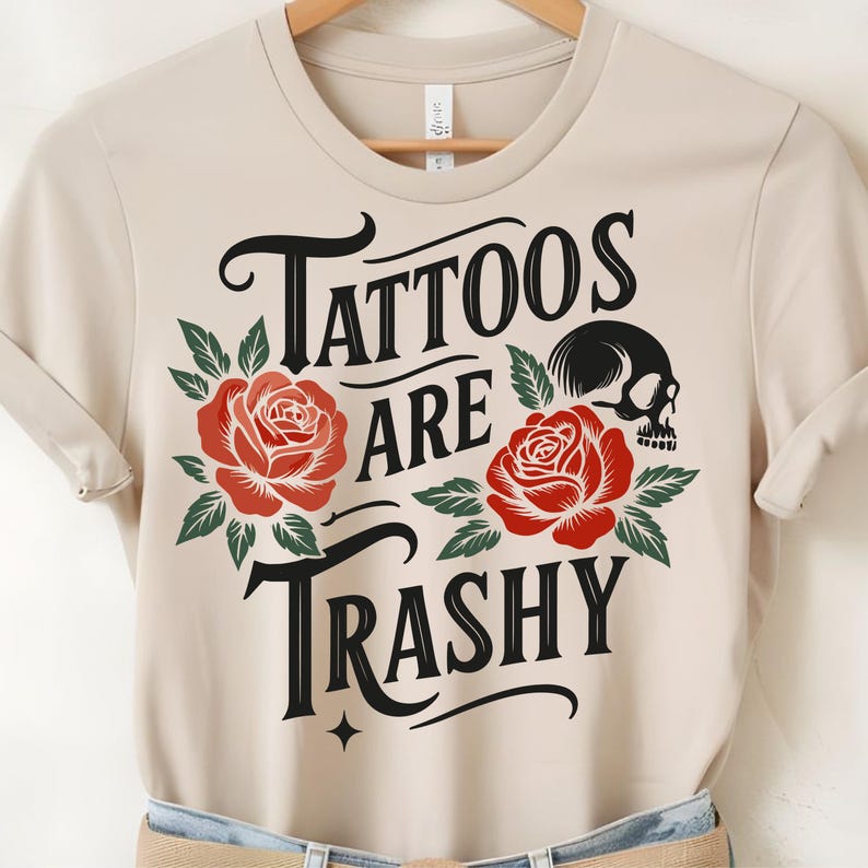 May include: Beige t-shirt with the text "Tattoos are Trashy" in black script, adorned with red roses and a skull graphic. The shirt is made of a soft material and has short sleeves.