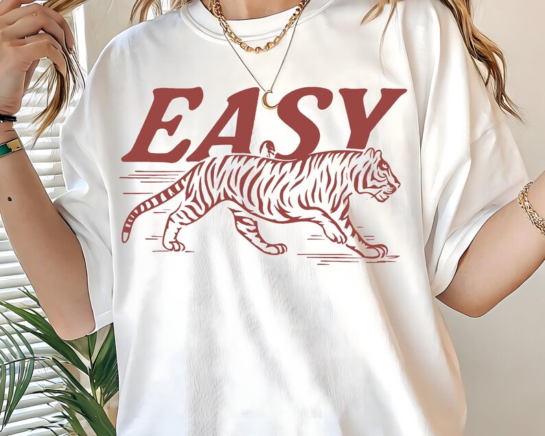 May include: White t-shirt with a red tiger graphic and the word "EASY" in a bold, serif font. The tiger is depicted in a walking pose with detailed stripes. The shirt is a casual, oversized style.