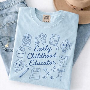 May include: Light blue t-shirt with a graphic design for an early childhood educator. The design includes a calculator, rainbow, notebook, lightbulb, apple, globe, pencil, and other school-related icons. The words "Early Childhood Educator" are printed in a dark blue script.
