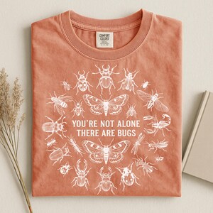 Op de afbeelding: Koraalrood T-shirt met ronde hals en een witte afbeelding van diverse insecten in een cirkel. De tekst "YOU'RE NOT ALONE THERE ARE BUGS" staat in het midden. Het shirt is gemaakt van zacht materiaal.