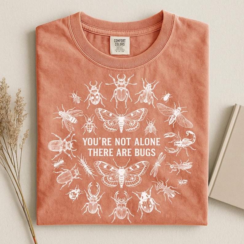 Op de afbeelding: Koraalrood T-shirt met ronde hals en een witte afbeelding van diverse insecten in een cirkel. De tekst "YOU'RE NOT ALONE THERE ARE BUGS" staat in het midden. Het shirt is gemaakt van zacht materiaal.