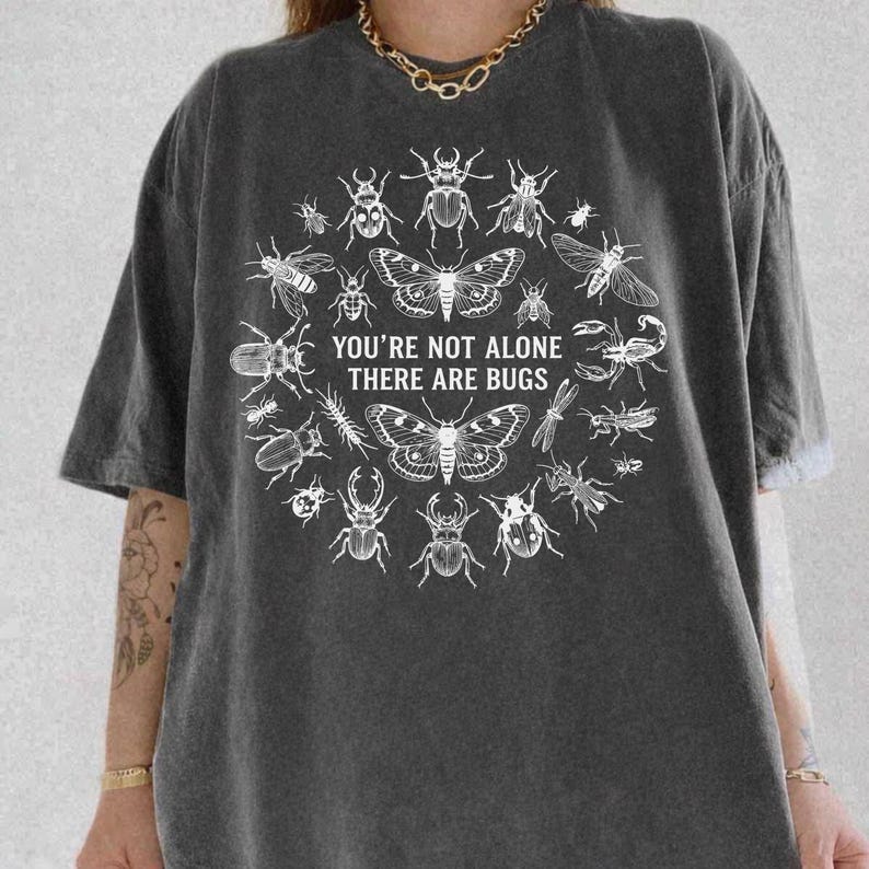 Op de afbeelding: Donkergrijs T-shirt met een cirkelvormig ontwerp van insecten, waaronder kevers, vlinders en een schorpioen. De tekst "YOU'RE NOT ALONE THERE ARE BUGS" staat in het midden. Het shirt heeft korte mouwen en een relaxte pasvorm.
