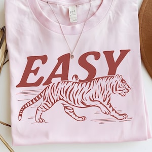 May include: A light pink t-shirt featuring the word "EASY" in a rust-colored font above a rust-colored tiger illustration. A delicate silver necklace with a small pendant is visible.