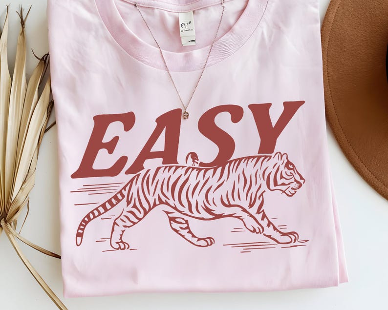 May include: A light pink t-shirt featuring the word "EASY" in a rust-colored font above a rust-colored tiger illustration. A delicate silver necklace with a small pendant is visible.