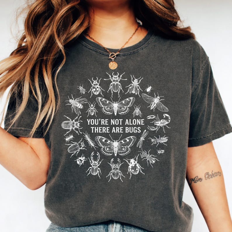 Op de afbeelding: Donkergrijs T-shirt met een witte insectenillustratie. Het ontwerp bevat vlinders, kevers en andere insecten in een cirkel. De tekst luidt "YOU'RE NOT ALONE THERE ARE BUGS". Het shirt is gemaakt van zacht materiaal.