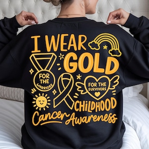 May include: Black sweatshirt with gold text and graphics. The design includes the words "I Wear Gold" and "Childhood Cancer Awareness." Graphics include a ribbon, heart with wings, medal, sun, and rainbow.