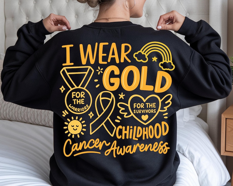 May include: Black sweatshirt with gold text and graphics. The design includes the words "I Wear Gold" and "Childhood Cancer Awareness." Graphics include a ribbon, heart with wings, medal, sun, and rainbow.