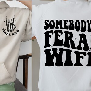 May include: A white hoodie with a black graphic of a skeleton hand making a rock and roll sign with the text "Feral Wife" around it. The back of the hoodie has the text "Somebody's Feral Wife" in black.