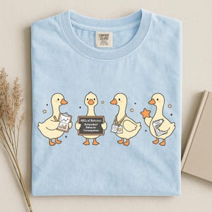 Op de afbeelding: Lichtblauw T-shirt met vier cartoon eenden die verschillende items vasthouden. Eén eend houdt een klembord vast, een ander een schoolbord met tekst, en anderen houden een ster en een document vast. Het shirt heeft een Comfort Colors label.