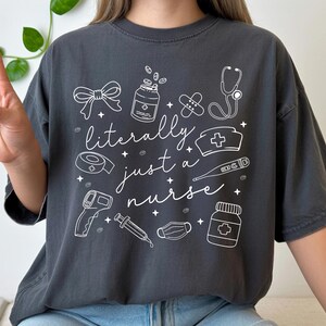 May include: Dark gray t-shirt with the phrase "literally just a nurse" and medical-themed illustrations in white. The design includes a stethoscope, pills, bandages, and a nurse's cap. The shirt is a casual, comfortable style.