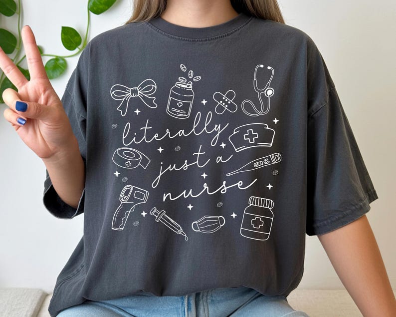 May include: Dark gray t-shirt with the phrase "literally just a nurse" and medical-themed illustrations in white. The design includes a stethoscope, pills, bandages, and a nurse's cap. The shirt is a casual, comfortable style.