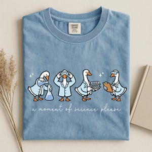 Puede incluir: Camiseta azul claro con una divertida ilustración de cuatro gansos con batas de laboratorio, realizando experimentos científicos. El texto "a moment of science please" está escrito debajo. Una prenda lúdica y educativa.