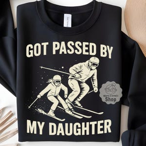 Op de afbeelding: Zwart sweatshirt met de witte tekst "GOT PASSED BY MY DAUGHTER". De afbeelding toont twee skiërs in witte skipakken en een skibril. De sweatshirt heeft een ronde hals en lange mouwen.