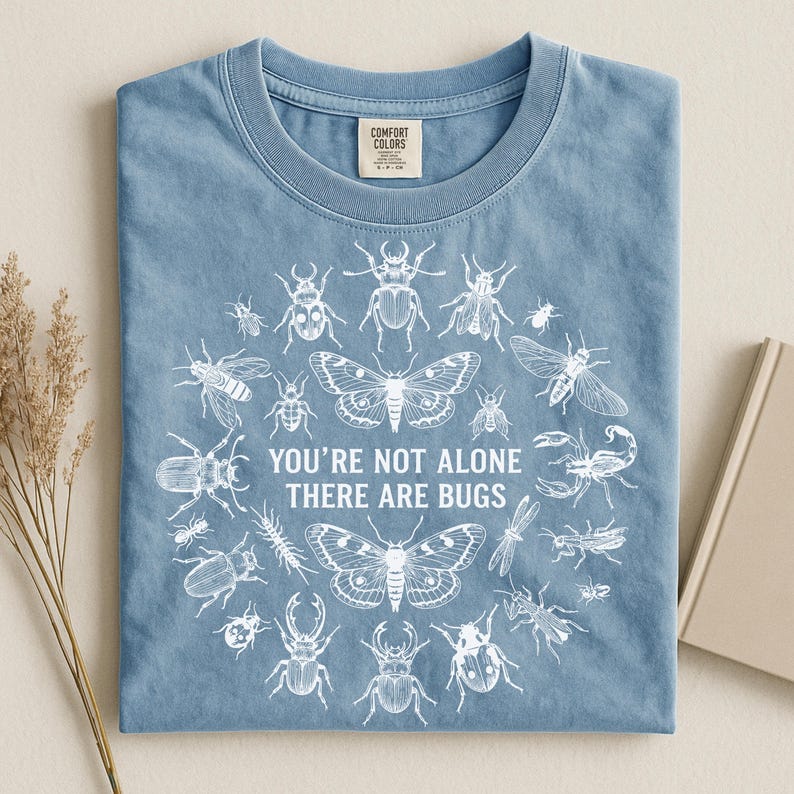 Op de afbeelding: Lichtblauw Comfort Colors T-shirt met een witte insectenillustratie. De tekst "YOU'RE NOT ALONE THERE ARE BUGS" staat in het midden. Het T-shirt is opgevouwen. Een takje gedroogd gras en een boek zijn zichtbaar.