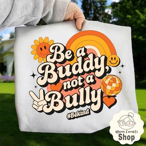 Peut inclure: Sac fourre-tout blanc avec un motif rétro. Le texte "Be a Buddy, not a Bully" est écrit dans une police groovy, avec une fleur souriante, un arc-en-ciel, une boule disco, des cœurs et un signe de paix. Le hashtag #Bekind est également inclus.