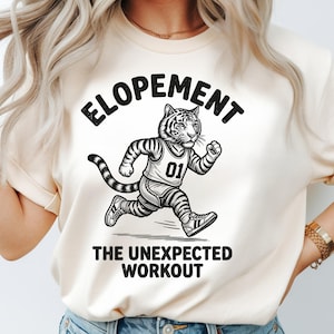 Pode incluir: Camiseta creme com um tigre correndo usando uma camisa com o número 01. A camisa tem as palavras "ELOPEMENT" acima do tigre e "THE UNEXPECTED WORKOUT" abaixo, tudo em texto preto.