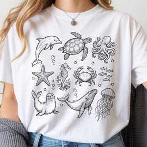 May include: White t-shirt featuring a black and white illustration of various sea creatures, including a dolphin, turtle, octopus, starfish, seahorse, crab, fish, seal, whale, and jellyfish. The design is a detailed line drawing.