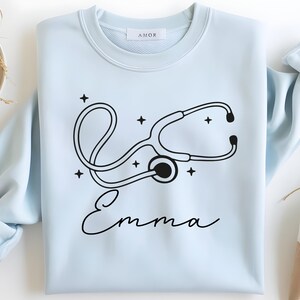 May include: A light blue sweatshirt featuring a black stethoscope design with the name "Emma" in cursive. The design includes stars around the stethoscope. The sweatshirt has a crew neck and long sleeves.