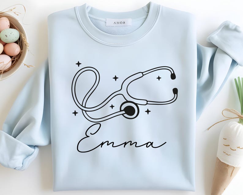 May include: A light blue sweatshirt featuring a black stethoscope design with the name "Emma" in cursive. The design includes stars around the stethoscope. The sweatshirt has a crew neck and long sleeves.