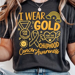 May include: Dark gray t-shirt with the text "I Wear Gold" and "Childhood Cancer Awareness" in yellow. The design includes a medal, ribbon, sun, rainbow, and heart with wings. The shirt's message supports childhood cancer awareness.