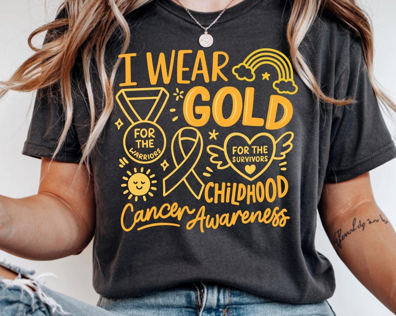 May include: Dark gray t-shirt with the text "I Wear Gold" and "Childhood Cancer Awareness" in yellow. The design includes a medal, ribbon, sun, rainbow, and heart with wings. The shirt's message supports childhood cancer awareness.