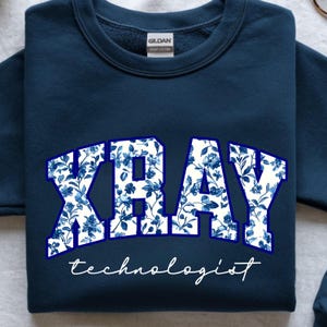 May include: Navy blue sweatshirt with the word "XRAY" in large, floral-patterned letters, and the word "technologist" in a cursive font. The floral pattern features blue flowers on a white background.