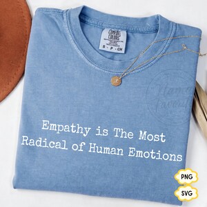 Może przedstawiać: Niebieski t-shirt z krótkim rękawem, okrągłym dekoltem i białym nadrukiem o treści, Empathy is The Most Radical of Human Emotions. Koszulka posiada metkę marki Comfort Colors przy wewnętrznej stronie kołnierza oraz złoty naszyjnik z wisiorkiem.