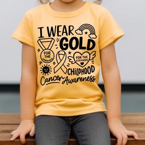 May include: A yellow t-shirt with the text "I Wear Gold for Childhood Cancer Awareness." The design includes a rainbow, ribbon, heart, and sun. The shirt is worn by a child.
