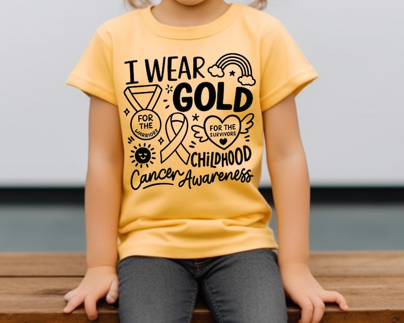 May include: A yellow t-shirt with the text "I Wear Gold for Childhood Cancer Awareness." The design includes a rainbow, ribbon, heart, and sun. The shirt is worn by a child.