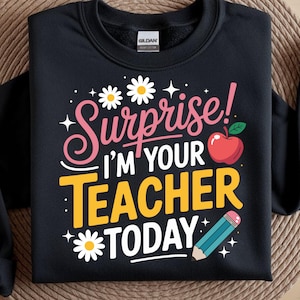 以下が含まれることがあります： ピンク、白、黄色の文字で「Surprise! I'M YOUR TEACHER TODAY」とプリントされた黒いクルーネックのスウェットシャツ。デザインには、2つの白いデイジー、緑の葉が付いた赤いリンゴ、ピンクの消しゴムが付いた青い鉛筆が描かれています。