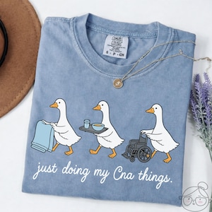 Puede incluir: Camiseta azul claro con un gráfico de tres patos de dibujos animados. Los patos se representan como cuidadores, llevando una toalla, una bandeja con comida y empujando una silla de ruedas. El texto "just doing my Cna things" está debajo.