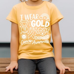 May include: A yellow t-shirt with the text "I Wear Gold for Childhood Cancer Awareness" in white. The shirt features a rainbow, ribbon, medal, sun, and heart graphics. The shirt is worn by a child.