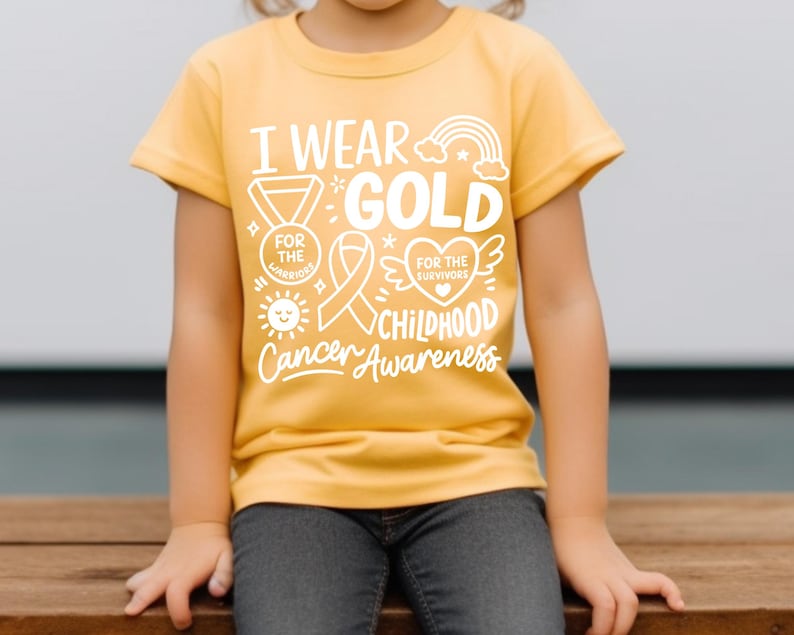 May include: A yellow t-shirt with the text "I Wear Gold for Childhood Cancer Awareness" in white. The shirt features a rainbow, ribbon, medal, sun, and heart graphics. The shirt is worn by a child.