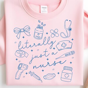 May include: A light pink sweatshirt with a blue graphic design. The design includes medical symbols such as a stethoscope, pills, and a nurse's hat, along with the text "literally just a nurse".