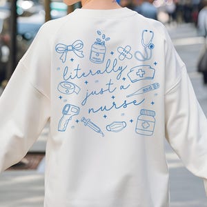 May include: Cream-colored sweatshirt with blue illustrations of medical tools and the phrase "literally just a nurse." The design includes a stethoscope, pills, bandages, and a thermometer. A casual, comfortable top for healthcare professionals.