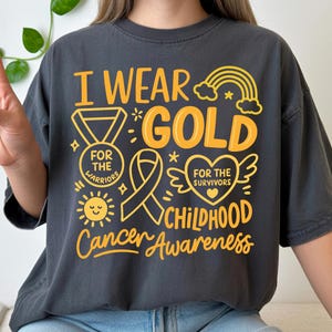 May include: Dark gray t-shirt with the text "I Wear Gold" in yellow, along with illustrations of a medal, ribbon, heart, rainbow, and sun. The shirt promotes Childhood Cancer Awareness.