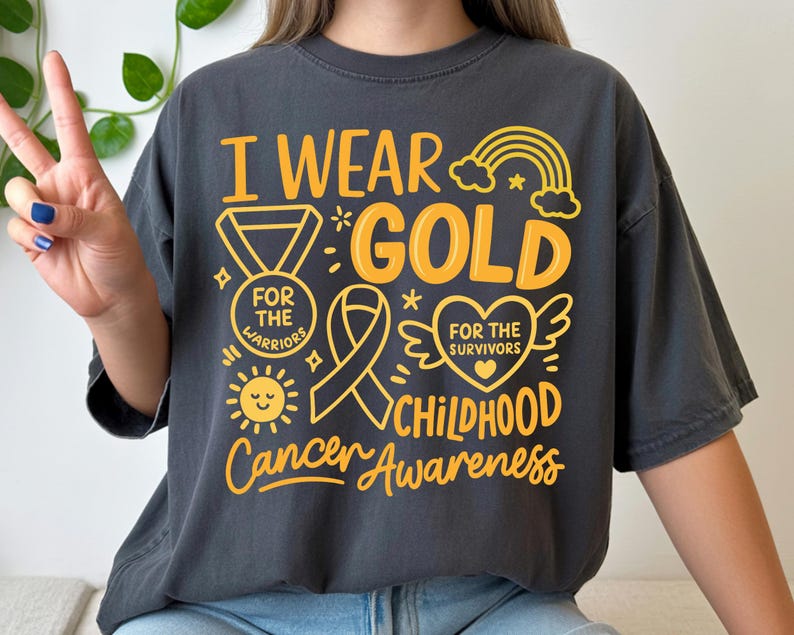 May include: Dark gray t-shirt with the text "I Wear Gold" in yellow, along with illustrations of a medal, ribbon, heart, rainbow, and sun. The shirt promotes Childhood Cancer Awareness.