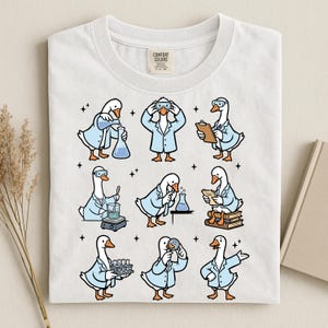 May include: White t-shirt with a repeating pattern of cartoon geese in lab coats, each engaged in different scientific activities. The geese are depicted with goggles and beakers, creating a playful science theme. The shirt has a Comfort Colors tag.