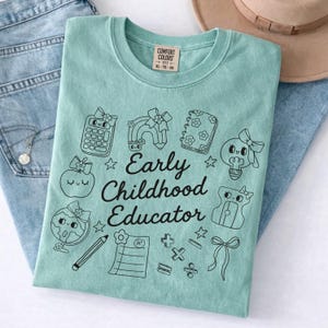 May include: A light teal Comfort Colors t-shirt with the words "Early Childhood Educator" and various black and white school-themed graphics. The shirt is folded on top of blue jeans.