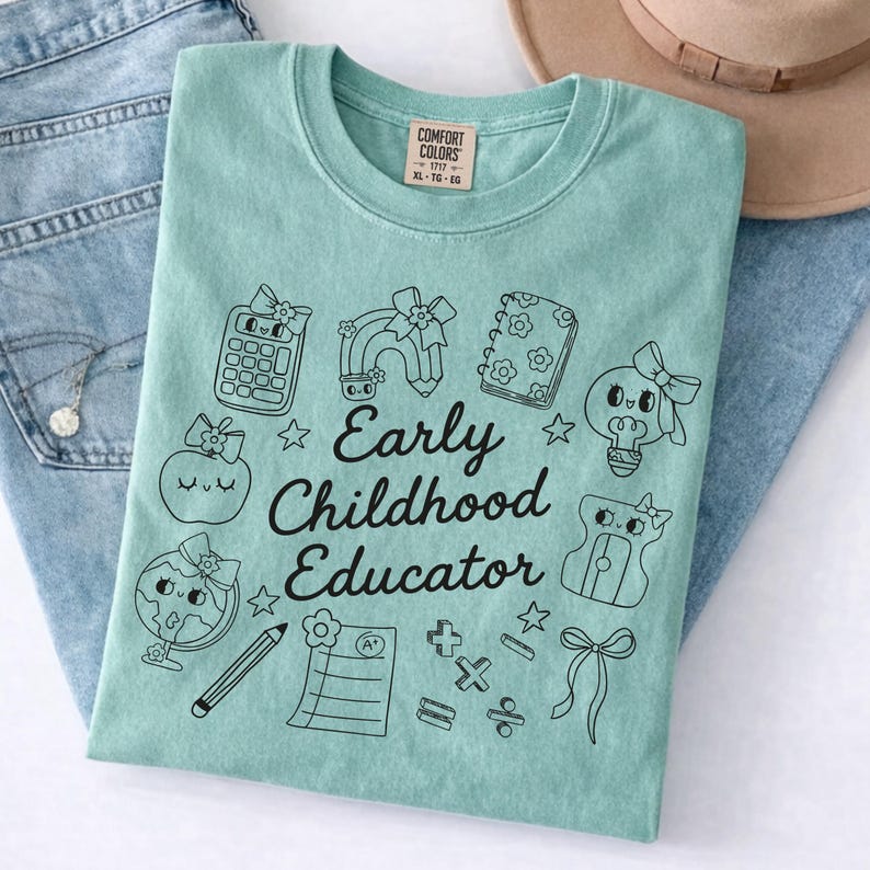 May include: A light teal Comfort Colors t-shirt with the words "Early Childhood Educator" and various black and white school-themed graphics. The shirt is folded on top of blue jeans.